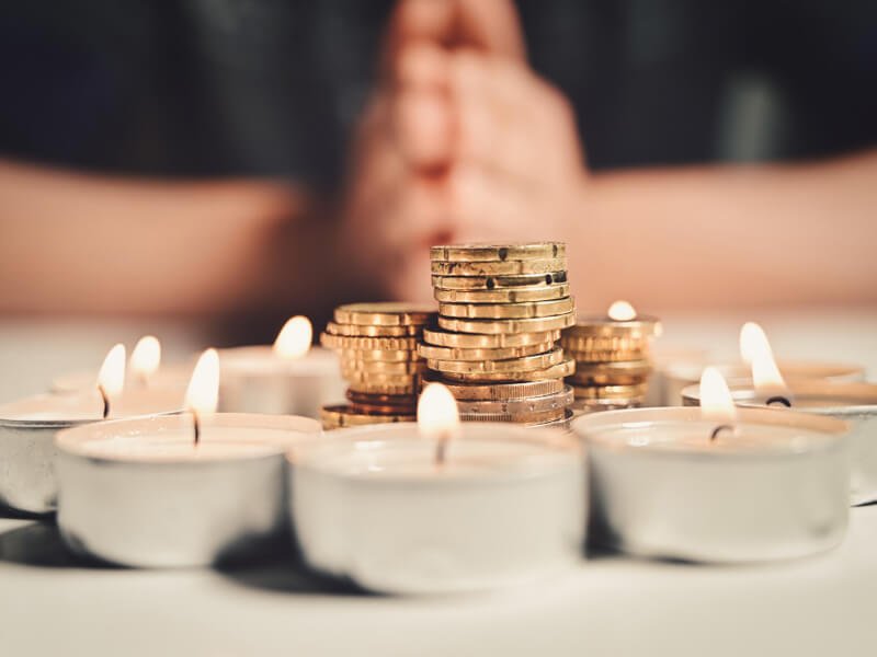 Hands of a man praying with a circle of burning candles with a stack of coins inside